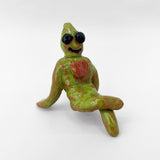 This ceramic sculpture portrays a whimsical green figure with a relaxed, laid-back pose, black eyes, a wide grin, and a red heart on its chest. The playful character exudes a quirky charm, combining elements of alien-like features with a lighthearted, friendly demeanor.