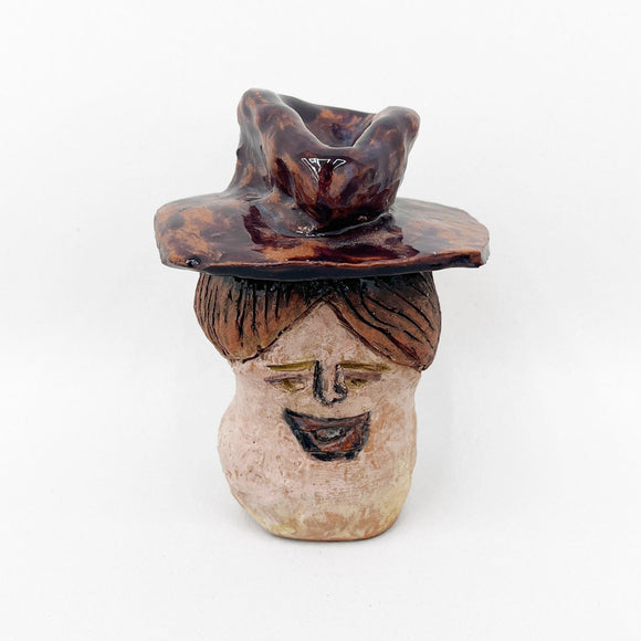 This ceramic sculpture depicts a stylized human face with exaggerated features, including a wide grin and closed eyes, topped by a large, textured hat with organic contours. The use of earthy tones and rough, uneven glazing techniques creates a rustic aesthetic, emphasizing the handmade quality and adding character to the figure's expression.