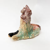 This ceramic sculpture presents a textured, abstract form that evokes the essence of a seated animal, perhaps a feline, with a slightly tilted head and alert posture. The earthy, muted colors, with touches of green, beige, and pink glazes, create a rough yet organic appearance, while the etched lines and glossy finish add depth and highlight the handcrafted quality of the piece.