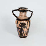 This ceramic vase, reminiscent of ancient Greek amphorae, features dual handles and is adorned with black-figure style imagery depicting silhouetted human and foliage motifs on a terracotta background. The contrast between the dark, expressive figures and the earthy clay body enhances the artifact's classical aesthetic, evoking themes of myth and antiquity.