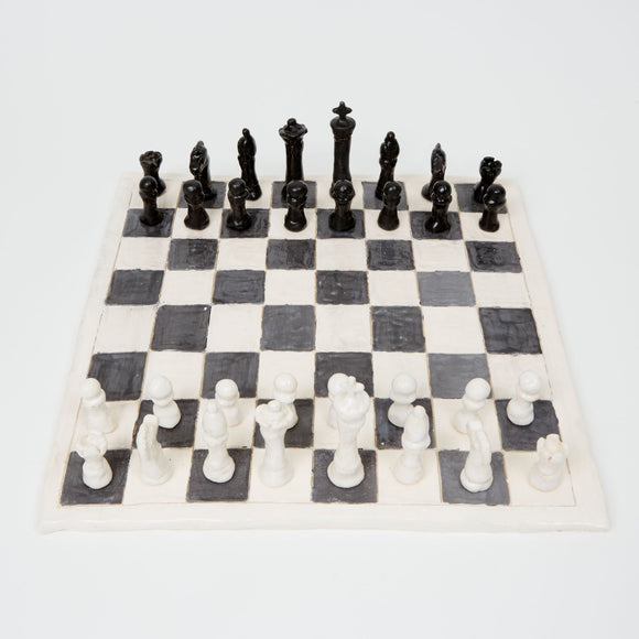 This ceramic chess set features handcrafted black and white pieces arranged on a classic checkered board, with each piece displaying a unique, slightly irregular form that emphasizes its artisanal nature. The contrast between the monochromatic palette and the tactile, imperfect surfaces of the pieces underscores a blend of functionality and aesthetic appeal, inviting reflection on the intersection of art and traditional game design.