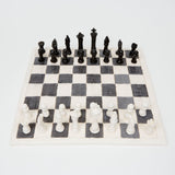 This ceramic chess set features handcrafted black and white pieces arranged on a classic checkered board, with each piece displaying a unique, slightly irregular form that emphasizes its artisanal nature. The contrast between the monochromatic palette and the tactile, imperfect surfaces of the pieces underscores a blend of functionality and aesthetic appeal, inviting reflection on the intersection of art and traditional game design.