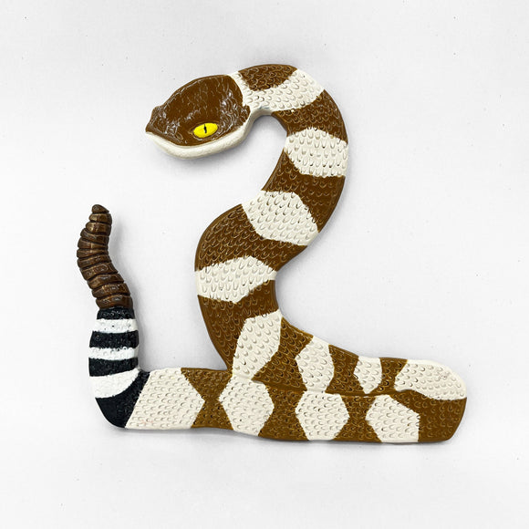 This sculpture depicts a stylized rattlesnake with a coiled body, alternating brown and white bands, and a textured surface that mimics scales. The exaggerated, bright yellow eye and segmented rattle emphasize both the snake's natural features and the artist's attention to detail, creating a vivid representation that balances realism with decorative abstraction.