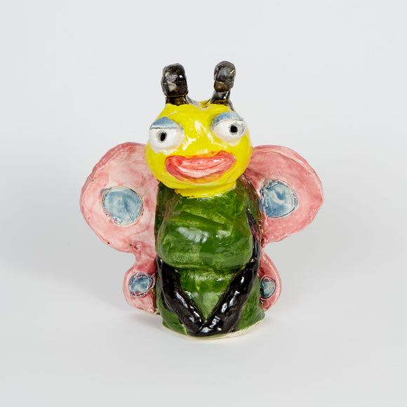 This ceramic sculpture depicts a whimsical insect-like figure with anthropomorphic features, including large eyes, bright pink lips, and prominent yellow and green coloration, accented by stylized pink wings with blue spots. The exaggerated facial features and glossy finish contribute to a playful, almost surreal quality, blending elements of realism and fantasy to represent a bug-like character.