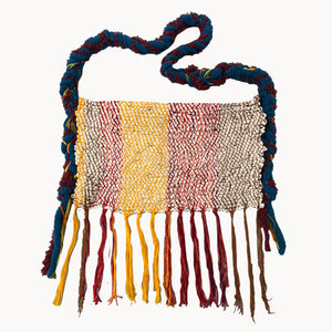 This handcrafted textile piece features a tightly woven, multicolored pattern in sections of cream, yellow, and red, accented by long, dangling fringe at the bottom. A thick, twisted strap in blue and maroon hues adds a sculptural element, enhancing the piece's tactile and visual complexity while suggesting both functionality and decorative intent.