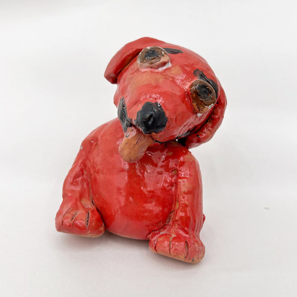 This ceramic sculpture depicts a stylized red dog with exaggerated features, such as large, black-patched eyes and a tilted head, giving it a playful, endearing expression. The glossy glaze enhances the vibrancy of the red hue, while subtle imperfections in form emphasize its handmade, expressive character.