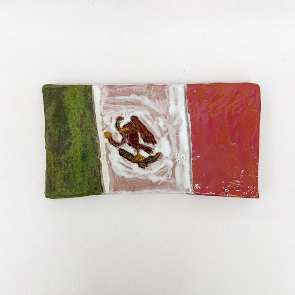 This ceramic piece represents the Mexican flag, featuring the traditional green, white, and red tricolor, with the national emblem of an eagle perched on a cactus in the center. The hand-painted glaze exhibits a textured finish, imparting a rustic quality that emphasizes the handcrafted nature of the object while celebrating national symbolism.