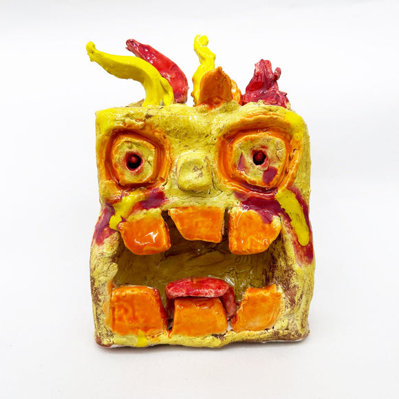 This ceramic sculpture portrays a stylized, fiery creature with exaggerated eyes, large square teeth, and flame-like protrusions, rendered in vivid shades of yellow, orange, and red. The textured surface and glossy finish enhance its expressive, monstrous quality, evoking themes of intensity and playfulness through a blend of abstract and anthropomorphic elements.