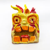 This ceramic sculpture portrays a stylized, fiery creature with exaggerated eyes, large square teeth, and flame-like protrusions, rendered in vivid shades of yellow, orange, and red. The textured surface and glossy finish enhance its expressive, monstrous quality, evoking themes of intensity and playfulness through a blend of abstract and anthropomorphic elements.