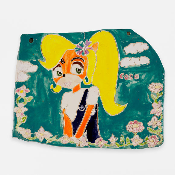 ‘Coco Bandicoot Cherry Blossom’ is a glazed ceramic piece that measures 8.5 by 10in. It features Coco Bandicoot, the younger sister of Crash Bandicoot, from the video game series with the same name. In this glazed ceramic piece, Coco stands in front of a sea green background with white clouds in the distance behind her. There is a string of green stems and pink flowers at the bottom of the piece. The name ‘Coco’ is written in pink paint. A multi-colored flower rests in Coco’s blonde hair.