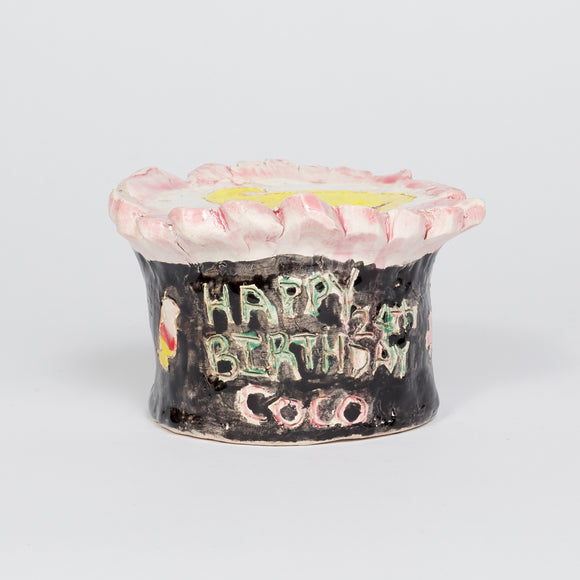 ‘Coco’s Birthday Cake’ is a glazed ceramic piece that measures 3 by 4.5 by 4.5in. It displays the words ‘Happy Birthday Coco’ in green and pink writing and has Coco Bandicoot’s face on top of the cake. The cake is cylindrical in shape and is decorated with white, pink, and black frosting. 