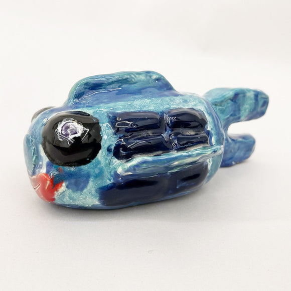 This ceramic sculpture depicts a stylized fish form, rendered in shades of blue with glossy, textured details that suggest scales and fins. The piece’s exaggerated features, such as a large eye and a subtly open mouth, lend it a whimsical quality, while the polished glaze enhances its aquatic theme and tactile appeal.