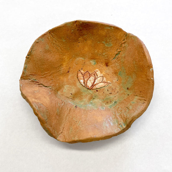 This ceramic bowl features an organic, irregular form with earthy tones of reddish-brown and subtle green patina, lending it a natural, handcrafted aesthetic. At its center, a delicate lotus flower design is etched in a slightly contrasting color, enhancing the piece’s meditative quality and evoking themes of tranquility and simplicity.