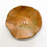 This ceramic bowl features an organic, irregular form with earthy tones of reddish-brown and subtle green patina, lending it a natural, handcrafted aesthetic. At its center, a delicate lotus flower design is etched in a slightly contrasting color, enhancing the piece’s meditative quality and evoking themes of tranquility and simplicity.