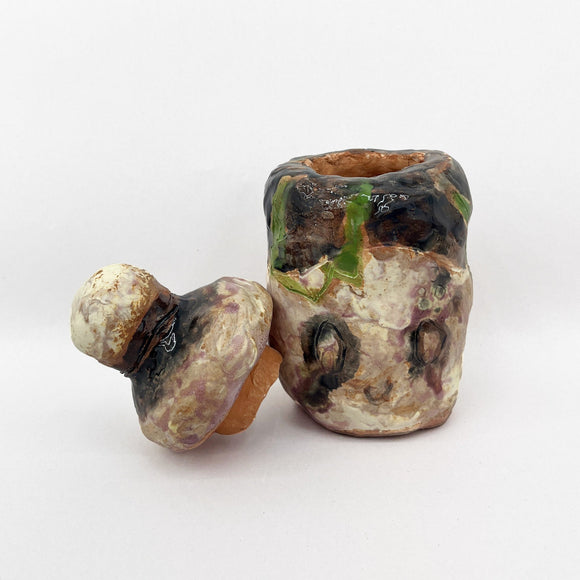 This ceramic artwork features a roughly textured container with an uneven, organic shape and a matching lid. The surface is covered in earthy tones of green, brown, and purple, giving it a natural, weathered appearance.