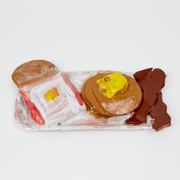 This ceramic sculpture depicts a stylized breakfast scene with pancakes topped with butter, toast, and strips of bacon arranged on a rectangular platter. The rough, hand-built forms and earthy, muted glazes lend the piece a whimsical, almost nostalgic quality, transforming an everyday meal into a playful exploration of texture and color.