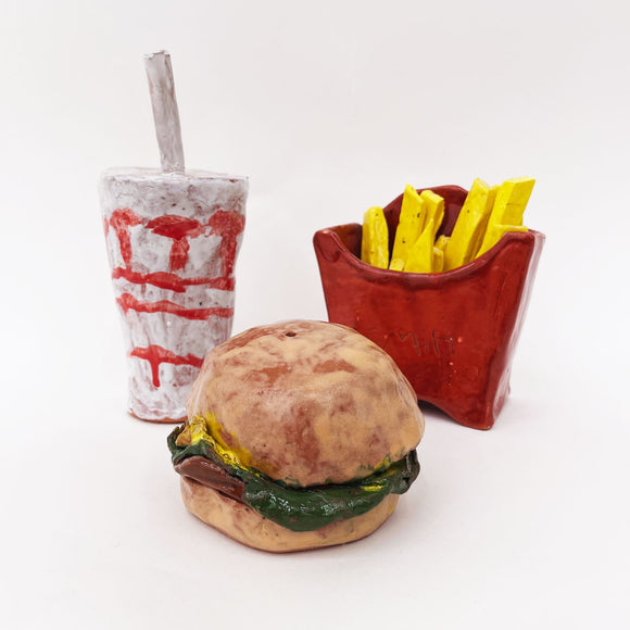 This ceramic piece depicts a classic fast-food meal with a burger, fries in a red container, and a drink cup, rendered in a playful and stylized manner. The rough, handmade textures and vibrant glazes capture the essence of each item, transforming familiar, everyday objects into a whimsical, sculptural representation of pop culture iconography.
