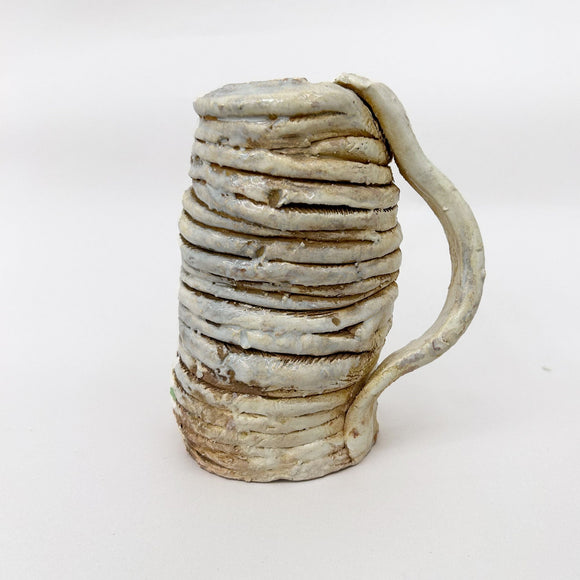 This ceramic piece is a textured mug constructed from coiled clay, with an earthy, uneven surface that emphasizes its handmade quality. The subtle, muted glaze highlights the organic, layered form, evoking a sense of rustic simplicity and connection to traditional pottery techniques.