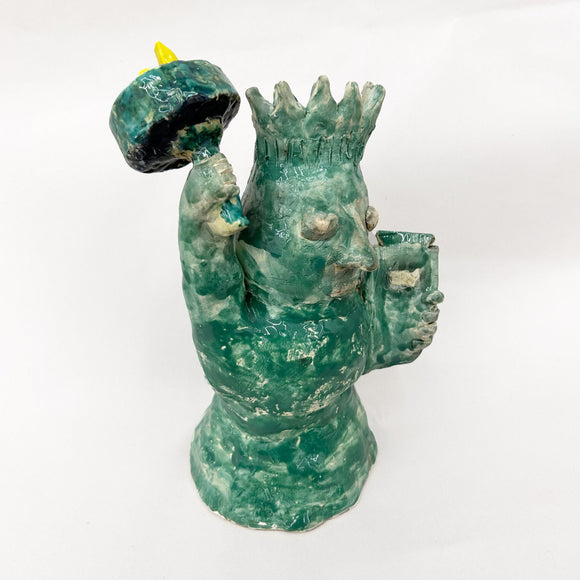 This ceramic sculpture portrays a stylized, whimsical figure reminiscent of the Statue of Liberty, with a textured green glaze and exaggerated facial features. Holding a torch and a tablet, the piece blends playful abstraction with iconic symbolism, offering an imaginative interpretation of a well-known monument.