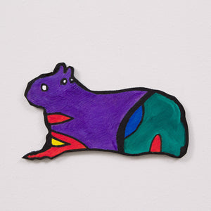 This 6 by 11 by ½-inch wood sculpture by Hugo Rocha portrays a stylized creature that resembles a capybara or guinea pig. The creature is painted in bright, bold colors: its main body is purple, its hindquarters are green with blue and red accents, and there are splashes of orange and red on its legs. The creature has a simple, cartoon-like face with white eyes and a small, friendly expression.