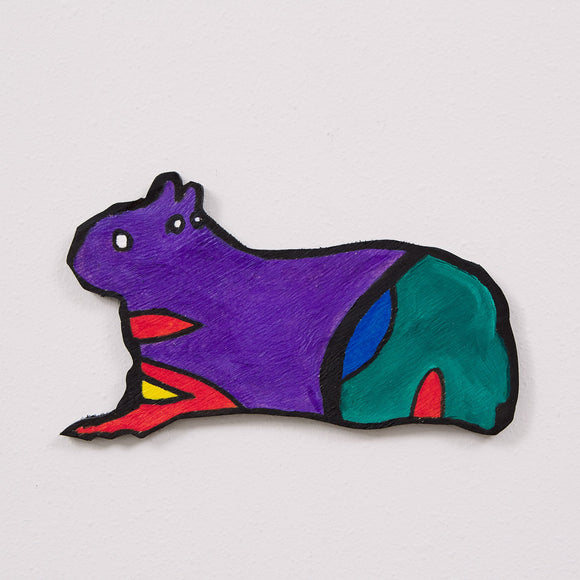 This 6 by 11 by ½-inch wood sculpture by Hugo Rocha portrays a stylized creature that resembles a capybara or guinea pig. The creature is painted in bright, bold colors: its main body is purple, its hindquarters are green with blue and red accents, and there are splashes of orange and red on its legs. The creature has a simple, cartoon-like face with white eyes and a small, friendly expression.