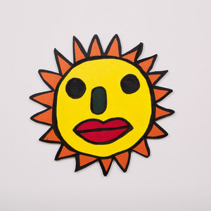 This 20 by 20 by 1/4th-inch wood sculpture by Hugo Rocha depicts a stylized sun with a human face. The sun's round, yellow face features bold, black eyes, a green nose, and red lips, giving it a playful and expressive character. The sun's rays are represented with orange pointed triangles that surround the face. The piece is outlined in black, emphasizing its vibrant colors and whimsical design.