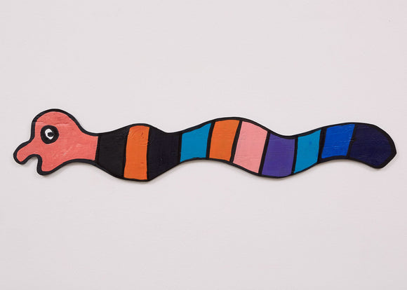 This 6 by 36 ½ by ½-inch wood sculpture by Hugo Rocha depicts a graphic, snake-like creature. The elongated body is segmented into colorful sections, including dark green, orange, pink, purple, blue, and dark blue. The head, painted a light red with a black outline, features a simple face with a large, expressive eye and a playful open mouth. The sculpture's vibrant, contrasting colors and smooth, wavy shape create a dynamic and cheerful character.
