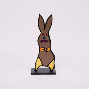 This 18 by 6 by ½-inch wood sculpture by Hugo Rocha depicts a stylized rabbit. The rabbit is painted brown with bright yellow accents on its paws and feet. Its face features a blue tear-shaped nose, red eyes, and pink lips, all outlined in black. The rabbit stands on a black rectangular base, giving it stability. The ears are long and pointed, adding to the curved shape of the rabbit.
