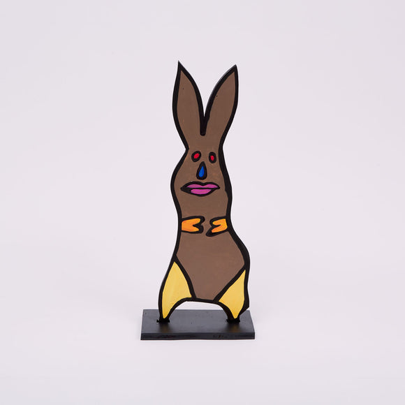 This 18 by 6 by ½-inch wood sculpture by Hugo Rocha depicts a stylized rabbit. The rabbit is painted brown with bright yellow accents on its paws and feet. Its face features a blue tear-shaped nose, red eyes, and pink lips, all outlined in black. The rabbit stands on a black rectangular base, giving it stability. The ears are long and pointed, adding to the curved shape of the rabbit.