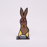 This 18 by 6 by ½-inch wood sculpture by Hugo Rocha depicts a stylized rabbit. The rabbit is painted brown with bright yellow accents on its paws and feet. Its face features a blue tear-shaped nose, red eyes, and pink lips, all outlined in black. The rabbit stands on a black rectangular base, giving it stability. The ears are long and pointed, adding to the curved shape of the rabbit.