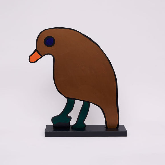 This 29 ½ by 23 by ½-inch wood sculpture by Hugo Rocha depicts a stylized bird. The bird's body is painted brown with a prominent orange beak and a deep blue eye. The legs are a dark green, contrasting with the body. The entire bird is outlined in black, emphasizing its shape. It stands on a black rectangular base, which supports its flat, two-dimensional form. The simplicity and bold use of color give the sculpture an abstract appearance.