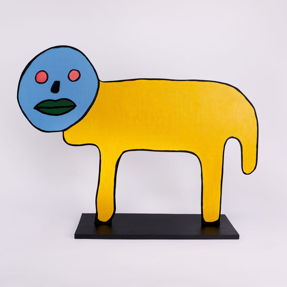 This wood sculpture by Hugo Rocha features a simple, abstract animal with a yellow body and a blue face. The face has two pink circular eyes, a green mouth, and a black nose, giving it a graphic expression. The body is rectangular with two straight legs, painted entirely in bright yellow. The sculpture is outlined in black, which contrasts with the vibrant colors and emphasizes its bold, cartoonish style. It stands on a black rectangular base.