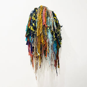 This textile artwork consists of a densely layered collection of multicolored yarns and fibers, cascading downward in a vibrant, textured mass. The varied materials, including twisted, knotted, and looped strands in earthy and bright hues, create a tactile, almost organic form that evokes natural elements like moss or seaweed, blending craft with abstract expression.