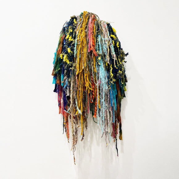 This textile artwork consists of a densely layered collection of multicolored yarns and fibers, cascading downward in a vibrant, textured mass. The varied materials, including twisted, knotted, and looped strands in earthy and bright hues, create a tactile, almost organic form that evokes natural elements like moss or seaweed, blending craft with abstract expression.