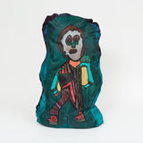 A painted cloth sculpture of a stylized figure with geometric black, pink, and teal patterns, large eyes, and a rectangular torso outlined in yellow and brown.