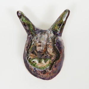 A dark, glossy ceramic mask with large, pointed ears and subtle green accents. The surface has textured lines and an expressive, abstract face.