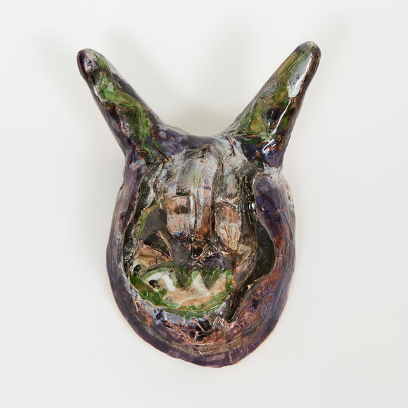 A dark, glossy ceramic mask with large, pointed ears and subtle green accents. The surface has textured lines and an expressive, abstract face.
