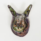 A dark, glossy ceramic mask with large, pointed ears and subtle green accents. The surface has textured lines and an expressive, abstract face.