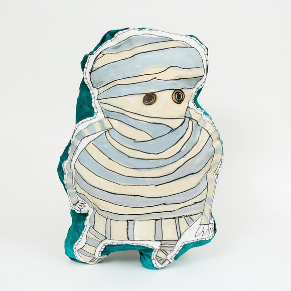 A soft sculpture painted with a wide-eyed, mummy-like character in pale blue and white stripes outlined in black, set against a teal background.