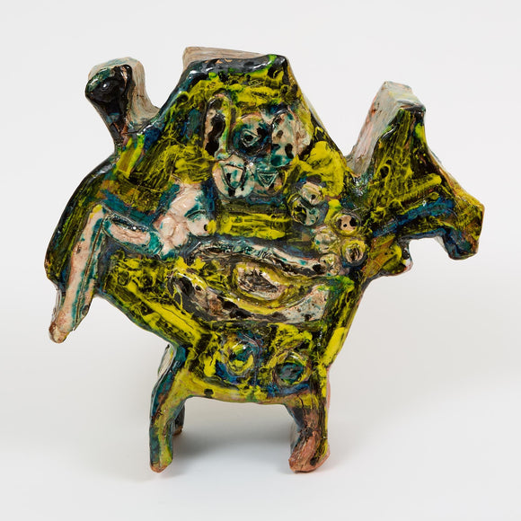 A ceramic sculpture in yellow, green, and black glazes, featuring a creature-like form with layered, textured surfaces and abstract facial features.