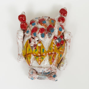 A wall-mounted ceramic work with a playful, mask-like form. The shiny surface features bright red, yellow, and blue details with bone-like protrusions.


