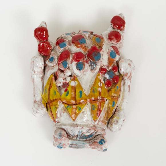 A wall-mounted ceramic work with a playful, mask-like form. The shiny surface features bright red, yellow, and blue details with bone-like protrusions.

