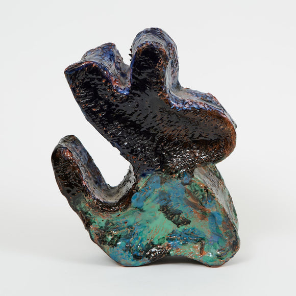 A glossy, abstract ceramic sculpture with angular, organic shapes—It resembles the shape of a seated dog. The dark glaze is accented by streaks of teal, green, and blue.