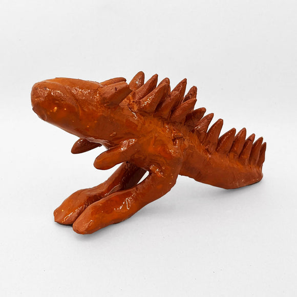 This ceramic sculpture represents a Carnotarus dinosaur with exaggerated, spiky dorsal fins and a glossy, earthy red glaze, evoking a prehistoric or mythical aesthetic. The textured surface and stylized anatomy give the figure a rugged, primal quality, merging elements of fantasy and natural form.