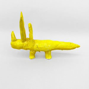 This ceramic sculpture features a bright yellow, abstracted Triceratops with two prominent upright spikes, resembling horns, and a long, elongated body. The textured surface and bold monochromatic glaze lend the piece a playful, surreal quality, blending simplicity with an imaginative, almost totemic presence.