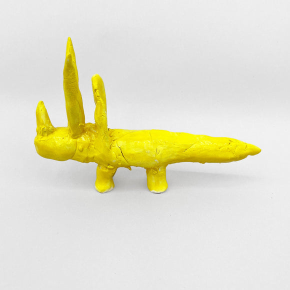 This ceramic sculpture features a bright yellow, abstracted Triceratops with two prominent upright spikes, resembling horns, and a long, elongated body. The textured surface and bold monochromatic glaze lend the piece a playful, surreal quality, blending simplicity with an imaginative, almost totemic presence.