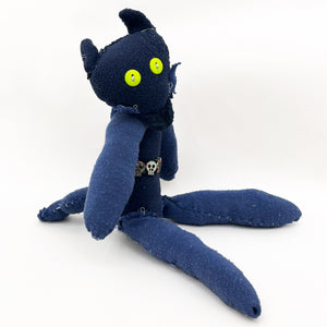 This handmade plush figure, constructed from dark blue fabric with visible stitching, combines simplicity with a touch of eerie charm through its bright green button eyes and skeletal belt accessory. The figure’s elongated limbs and rough, stitched-together texture evoke a sense of imperfection and individuality, suggesting a playful yet slightly gothic character.