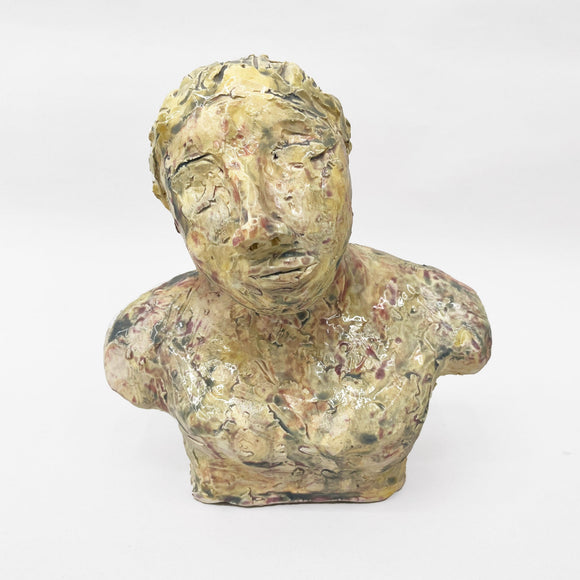 This ceramic bust portrays a figure with a contemplative and somewhat melancholic expression, rendered in a textured, earth-toned glaze that gives the surface a raw, organic quality. The detailing of the face, with its closed or averted eyes and tilted head, suggests a state of introspection or vulnerability, inviting viewers to ponder the emotional depth conveyed by the artist.