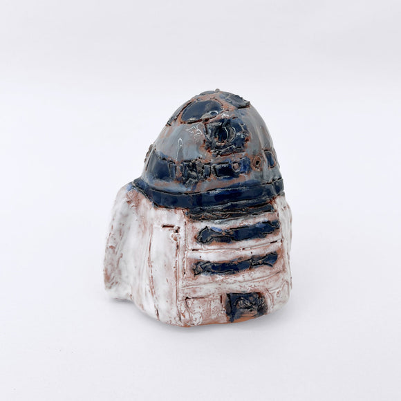 This ceramic sculpture is a stylized representation of the iconic Star Wars character R2-D2, capturing its distinctive domed top and cylindrical body with a hand-crafted, organic aesthetic. The surface is adorned with etched lines and glazes that reinterpret R2-D2’s familiar mechanical details through an artisanal, earthy approach, bridging pop culture with traditional ceramic techniques.