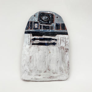 This ceramic relief sculpture reimagines R2-D2 with a flattened, almost minimalist interpretation, focusing on the droid’s iconic front panel details. The muted glazes and rough texture bring an organic quality to the mechanized subject, offering a tactile reinterpretation of the beloved character from Star Wars.